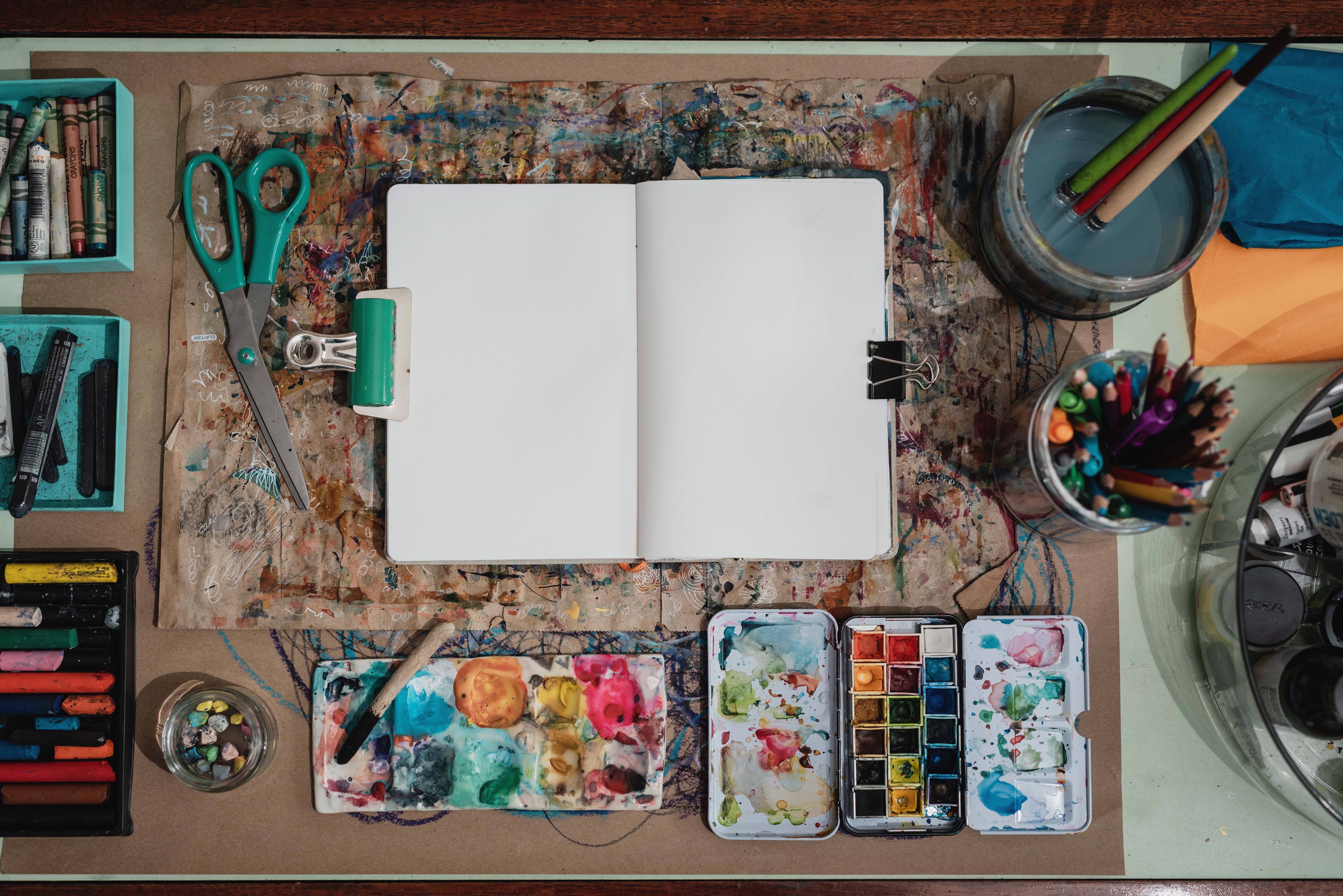 A overhead view of a blank notebook and various art supplies, including paints, pastels, and pencil crayons.